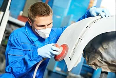 Механік шліфує білу деталь автомобіля для підготовки до фарбування.