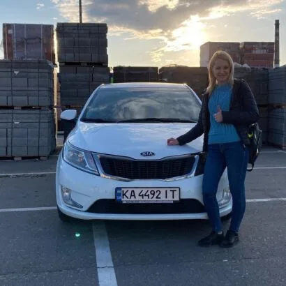 Жінка стоїть поруч із білим автомобілем на парковці.